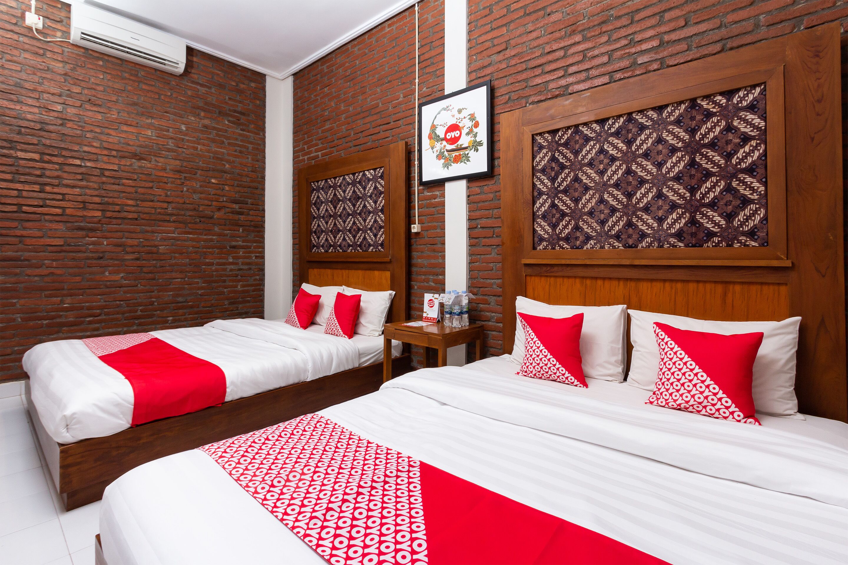 Habitación de hotel con dos camas, cada una con ropa de cama blanca y detalles rojos, una pared de ladrillo y una obra de arte enmarcada sobre las camas.