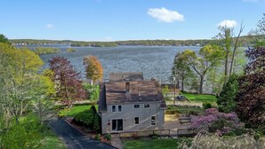 A two-story house with a front yard, a driveway, and a view of a lake. - Grammy's Lake House on Lake Pocotopaug (East Hampton)