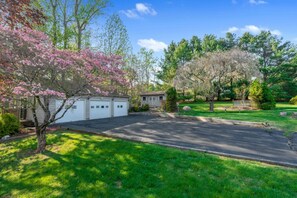 Une allée menant à une propriété résidentielle, avec un garage, une petite maison et une pelouse agrémentée d’arbres.