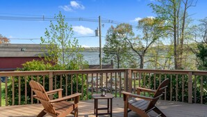 A wooden deck with two Adirondack chairs and a small table, overlooking a body of water and trees. - Grammy's Lake House on Lake Pocotopaug (East Hampton)