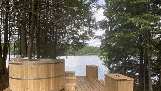 Puinen terassi, jossa on suuri puutynnyri, puiset portaat ja näkymä puilla ympäröidylle järvelle.