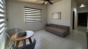 Ein Wohnzimmer mit Deckenventilator, einer Couch, einem Esstisch mit Stühlen und einem Fenster mit Jalousien.