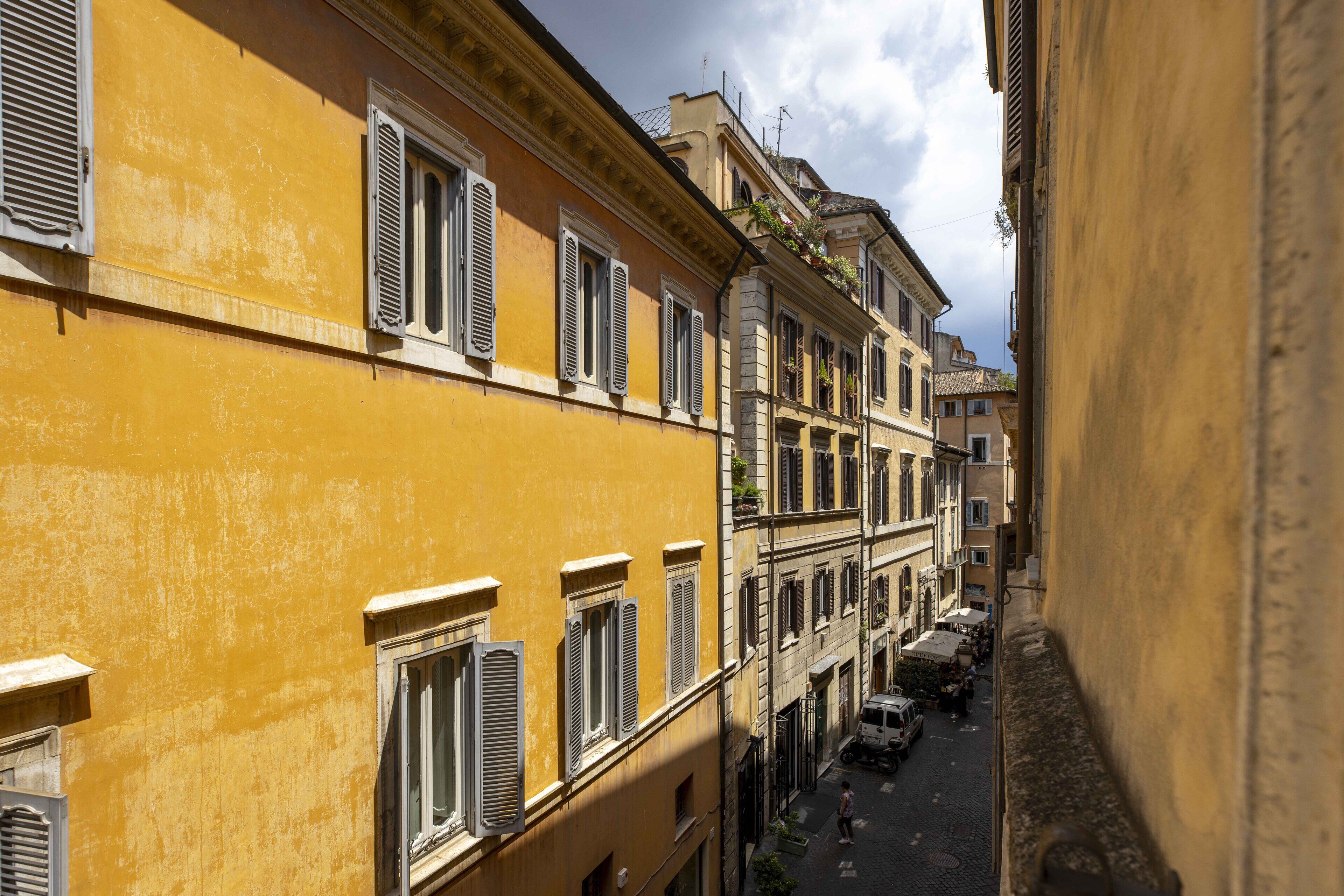 A narrow street lined with yellow buildings with shutters, a cobblestone road, and a parked vehicle.