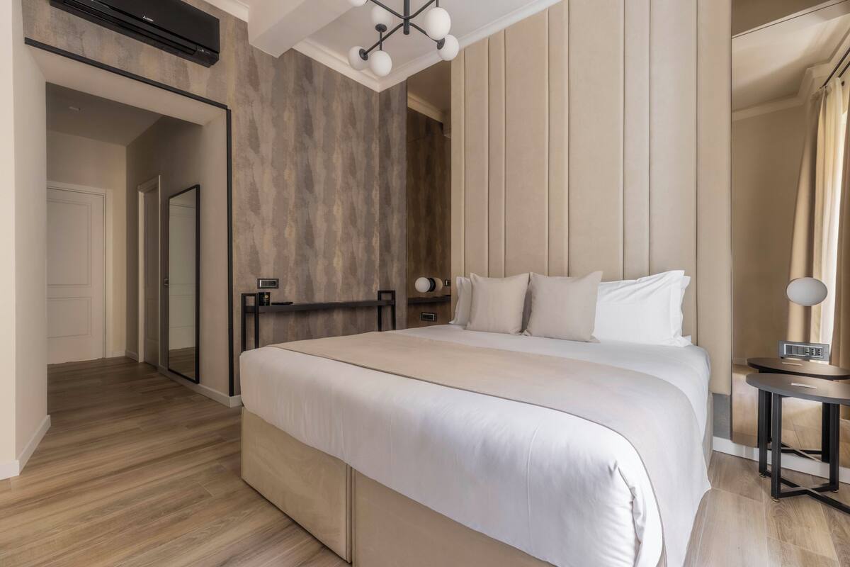 A modern hotel room with a large bed, a bedside table, a mirror, and a built-in wardrobe.
