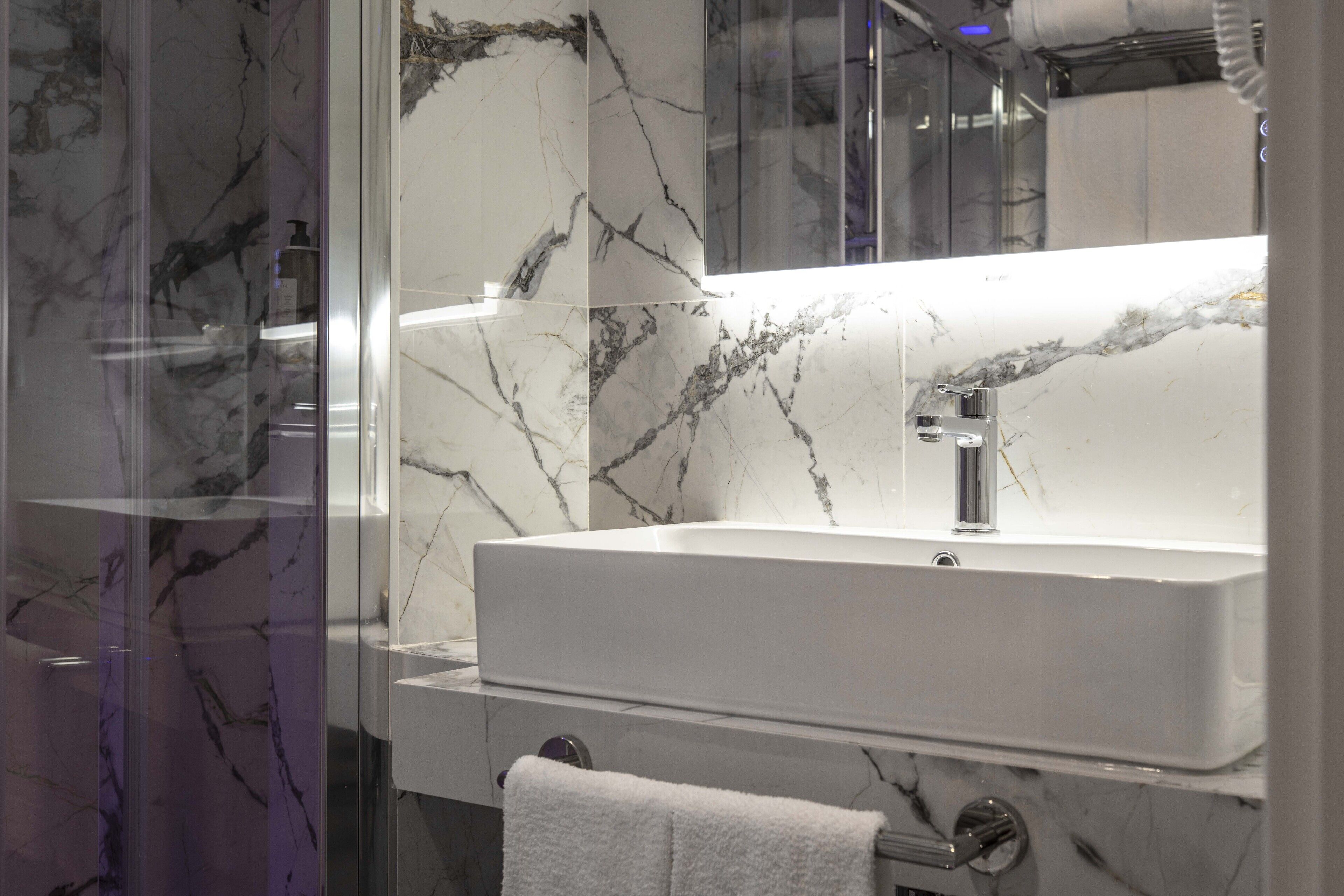 Une salle de bain avec un lavabo blanc, des murs en marbre et une cabine de douche en verre.