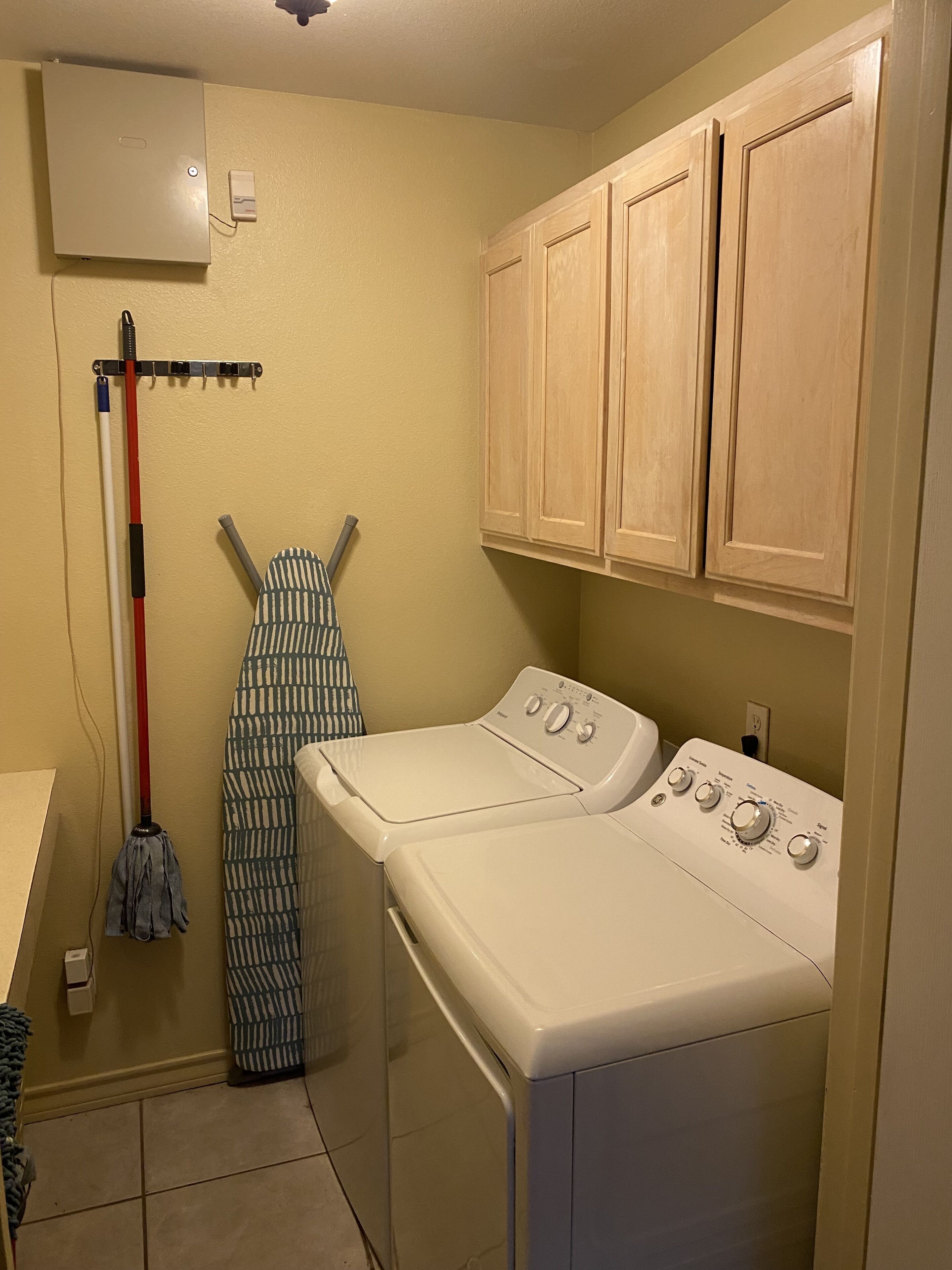 A laundry room with a washing machine, ironing board, and mops.