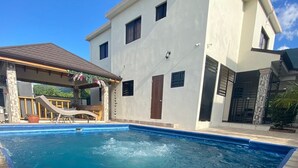 A rectangular swimming pool with a stone border, a covered patio area with seating, and a two-story building with a flat roof and a brown door. - Romantic VillaLunaCaridad (Jarabacoa)