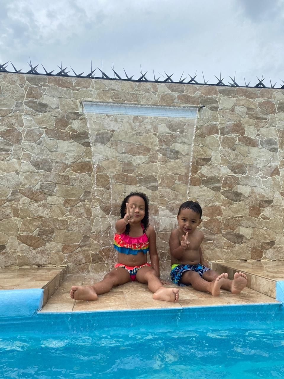 Zwei Kinder sitzen an einem Pool, im Hintergrund eine Steinmauer und ein Metallzaun.