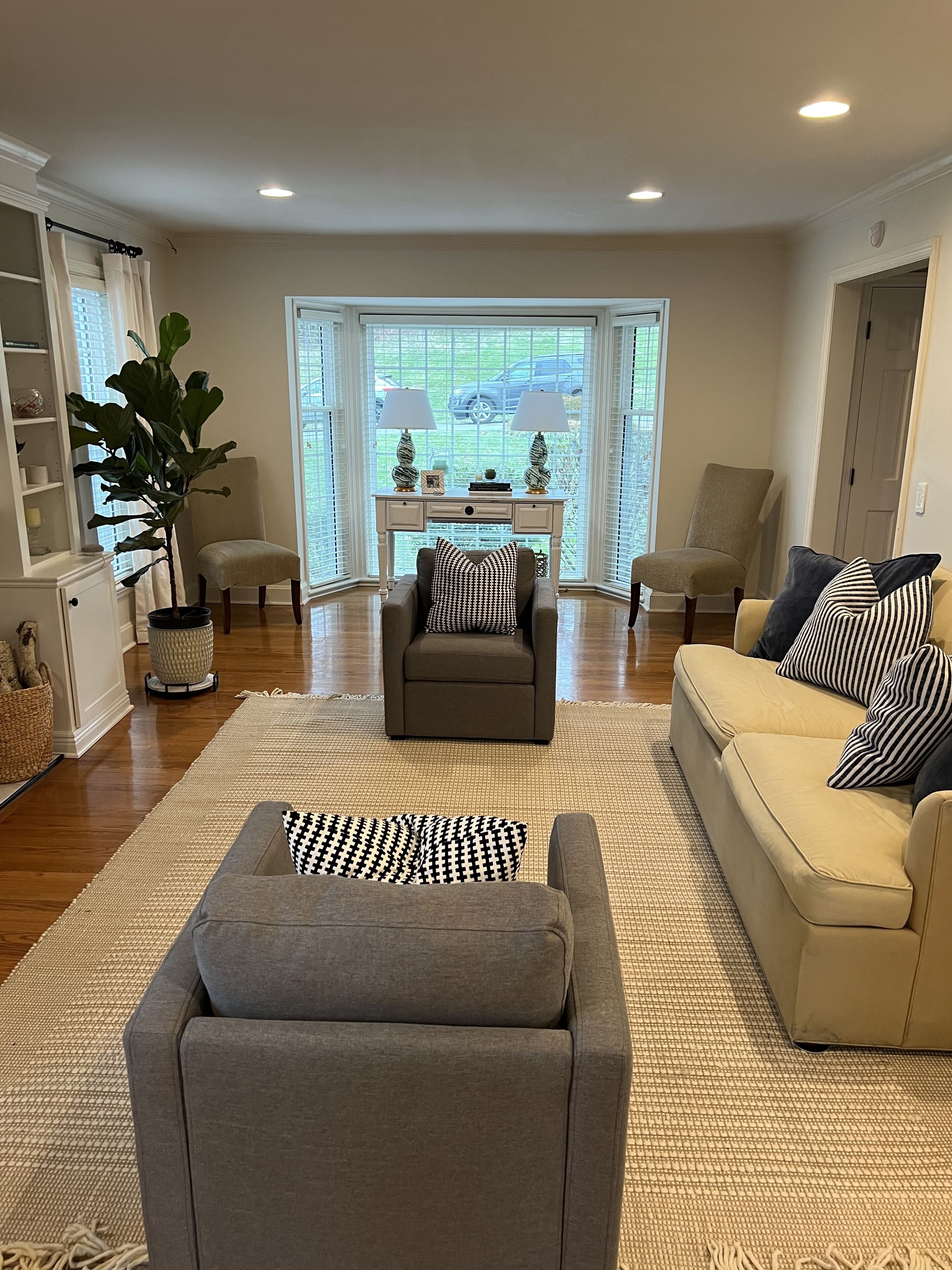 A living room with a grey sofa, a beige sofa, and a grey armchair. There is a large window with blinds, a white coffee table, and two lamps. A potted plant is on the left side.
