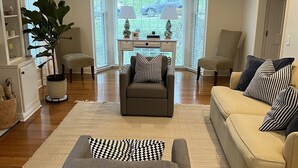 Ein Wohnzimmer mit grauem Sofa, beigem Sofa und grauem Sessel. Ein großes Fenster mit Jalousien, ein weißer Couchtisch und zwei Lampen sind vorhanden. Eine Topfpflanze steht links.