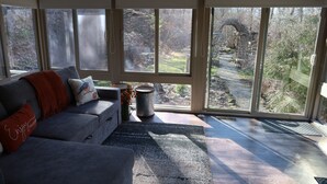 A modern living room with a gray sofa, a red throw blanket, and a view of a garden through large windows. - Whitetail Ridge Cabin ,Sleeps 10 (Loudonville)