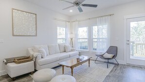 Uma sala de estar moderna com um sofá branco, uma mesa de centro e uma poltrona. Há um ventilador de teto, janelas amplas e vista para o exterior.