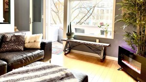 Ein Wohnzimmer mit einem Ledersofa, einem gemusterten Teppich, einem Fernseher und einem großen Fenster mit Blick auf Bäume und Gebäude.