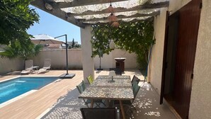A covered patio with a pool, a table with chairs, and a hanging light fixture. - Villa Port Camargue – Private pool, air conditioning, free parking (LE GRAU DU ROI)