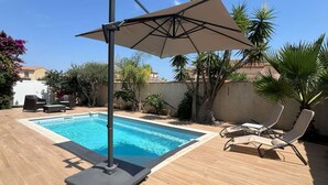 A swimming pool with a sun umbrella, lounge chairs, and a wooden deck. - Villa Port Camargue – Private pool, air conditioning, free parking (LE GRAU DU ROI)