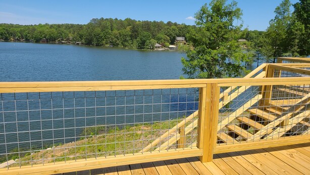 Une terrasse en bois avec une rambarde métallique donnant sur un lac, entourée d’arbres et d’un bâtiment situé au loin.