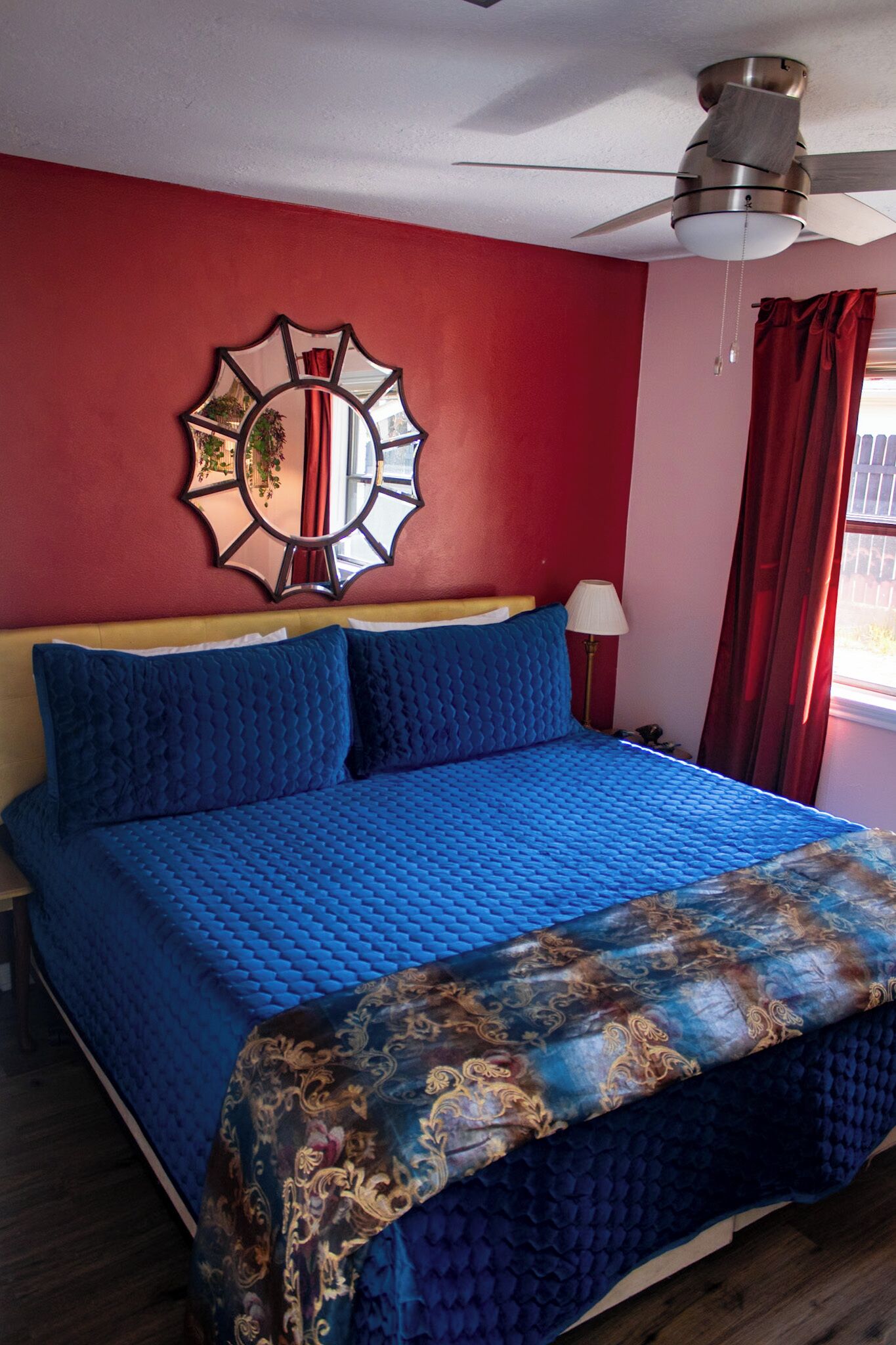 Un lit avec une couette bleue, un miroir décoratif et un ventilateur de plafond.