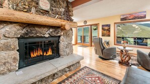 A stone fireplace with a fire, a stone shelf with a sign, and a view of mountains through large windows. - Divine Destiny Vacation Home at Windcliff (Estes Park)