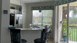 A kitchen with a central island, two bar stools, a refrigerator, and a window with blinds. - Pelican Sound Golf and River Club (Estero)