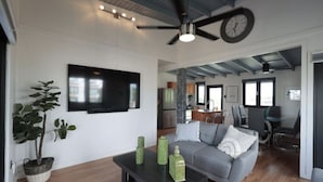 A modern living room with a grey sofa, a coffee table with decorative items, a large flat-screen TV, and a ceiling fan. - The Good Vibes House (Cabo Rojo)