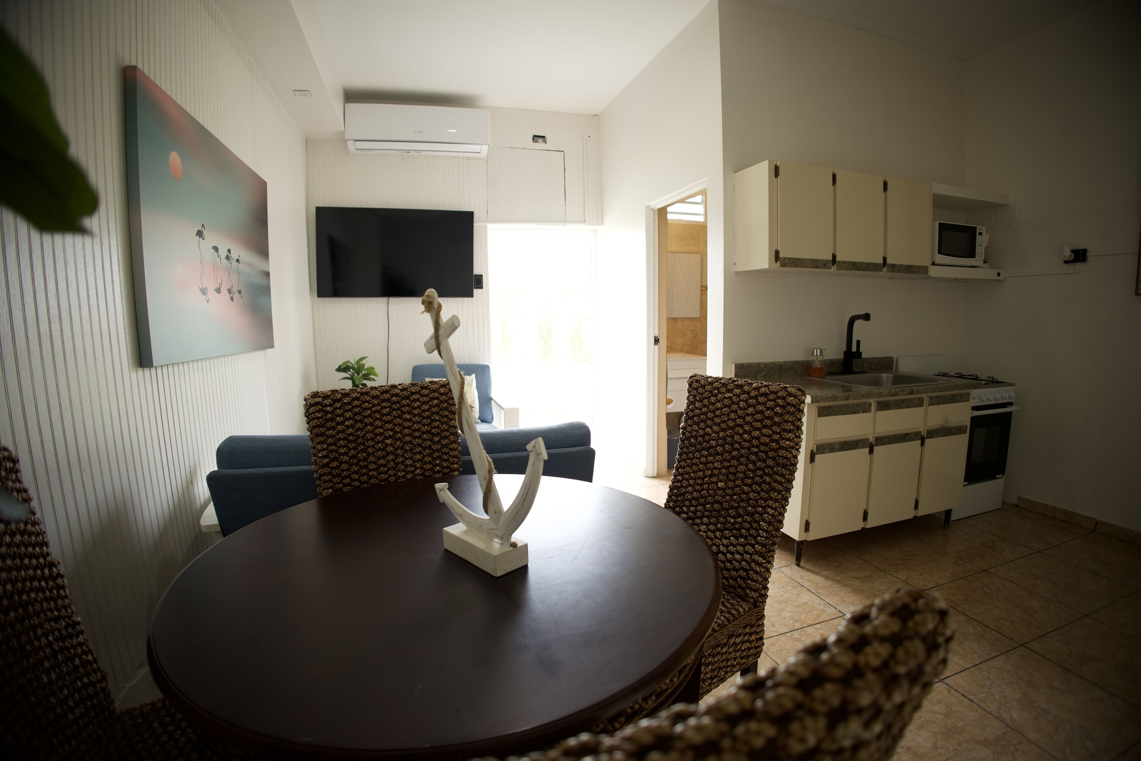 A modern living room with a round dining table, wicker chairs, a flat-screen TV mounted on the wall, and a kitchen area with white cabinets and a microwave.