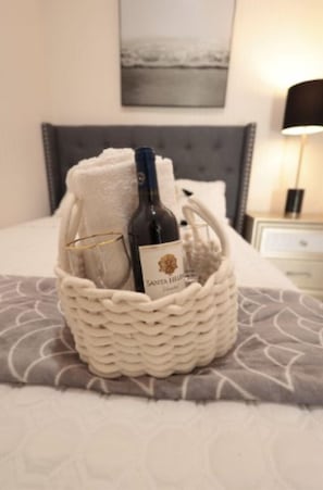 A bed with a grey patterned bedspread, a white woven basket containing a bottle of Santa Lucia wine, a glass, and a white towel. - The Good Vibes House (Cabo Rojo)