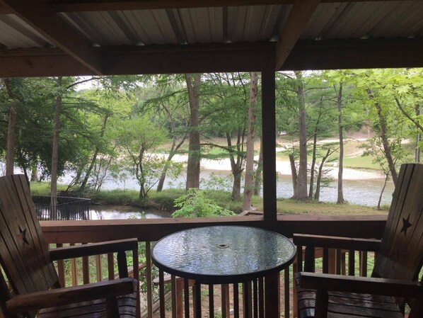 A wooden deck with a glass table and two chairs, overlooking a lake surrounded by trees.