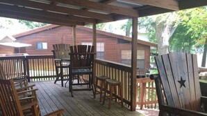 A covered wooden deck with several Adirondack chairs and a table, surrounded by a wooden fence. - Rio Cancion on Guadalupe Cabin 2 (New Braunfels)
