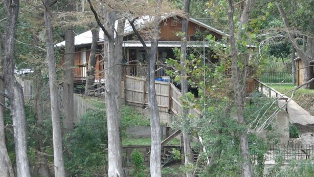 A wooden house with a deck, surrounded by trees and a body of water. - Rio Cancion on Guadalupe Cabin 2 (New Braunfels)