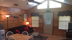 A cozy cabin interior with a wooden interior, a dining area with a table and chairs, a sofa with red cushions, a lamp, and a television. - Rio Cancion on Guadalupe Cabin 2 (New Braunfels)