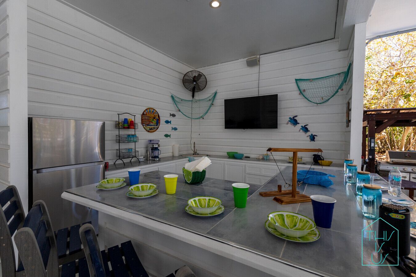A kitchen with a stainless steel refrigerator, a wooden table with various dishes and cups, a wall-mounted fan, and a television.