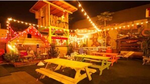 Un espacio al aire libre con mesas y bancos de madera, luces colgantes y un edificio con un letrero.