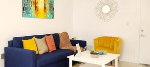 Un salon avec un canapé bleu, un fauteuil jaune, une table basse blanche et un tableau accroché au mur.