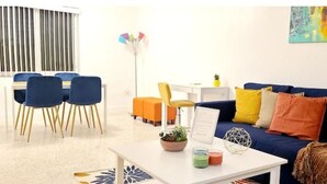Una sala de estar moderna con un sofá azul, sillones naranjas, una mesa de centro blanca con una planta y un cuadro en la pared.