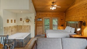A wooden cabin interior with a large bathtub, two beds, a dining area, and a ceiling fan. - Rustic Studio Cabin with King Bed and Jacuzzi Tub (Branson)