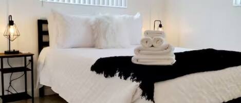 Un lit bien fait, avec du linge de lit blanc, un plaid noir et un ensemble de serviettes posées sur le lit. Il y a une petite table de chevet avec une lampe, et le sol de la pièce est recouvert d’un revêtement à motifs.