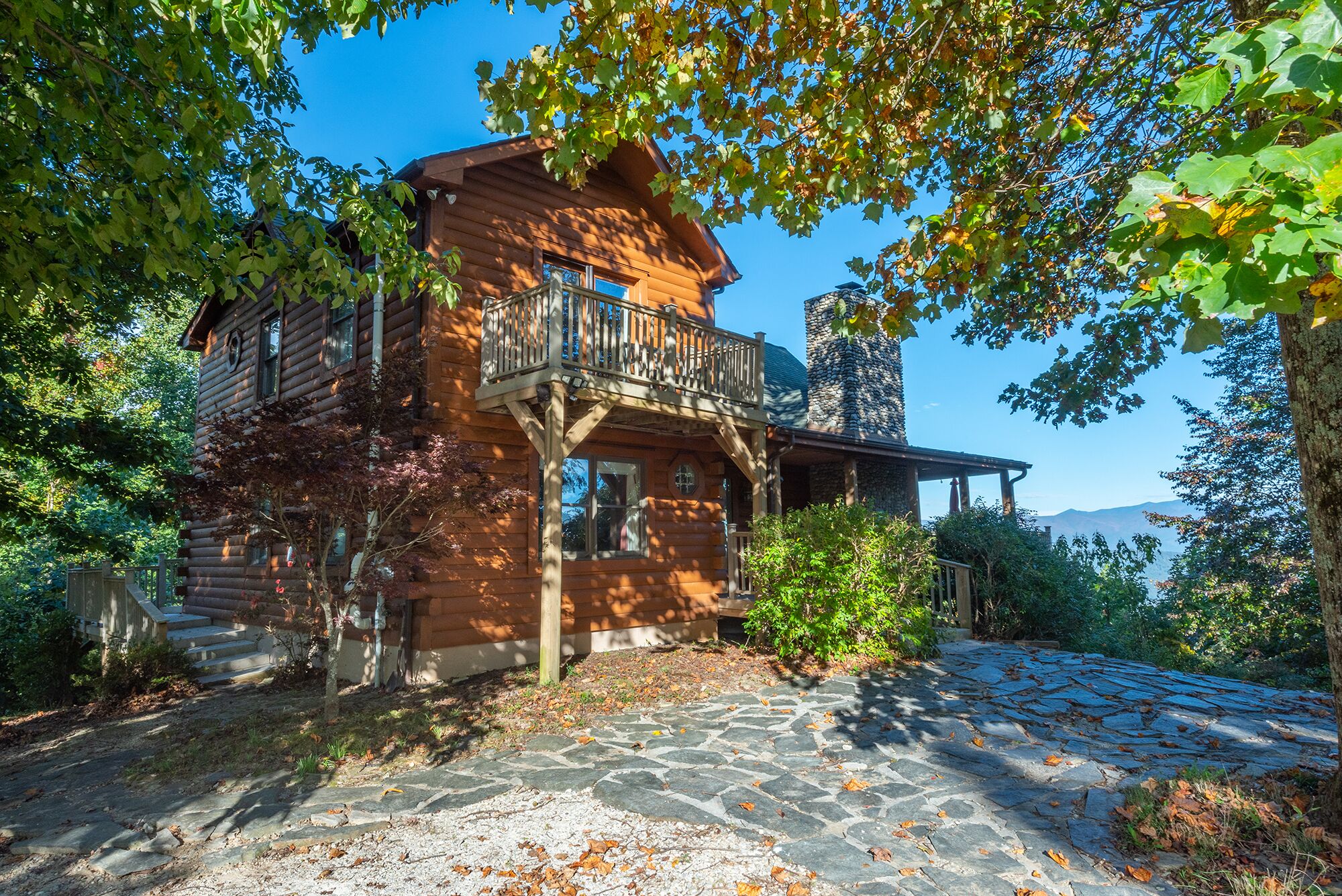 A log cabin with a balcony, stone pathway, and surrounding trees.