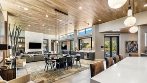 Un vaste espace de vie comprenant un grand canapé, une table à manger et des chaises. Il y a également une cuisine avec un grand îlot central et une table à manger. La pièce est dotée d’un haut plafond avec des poutres en bois et un éclairage encastré.