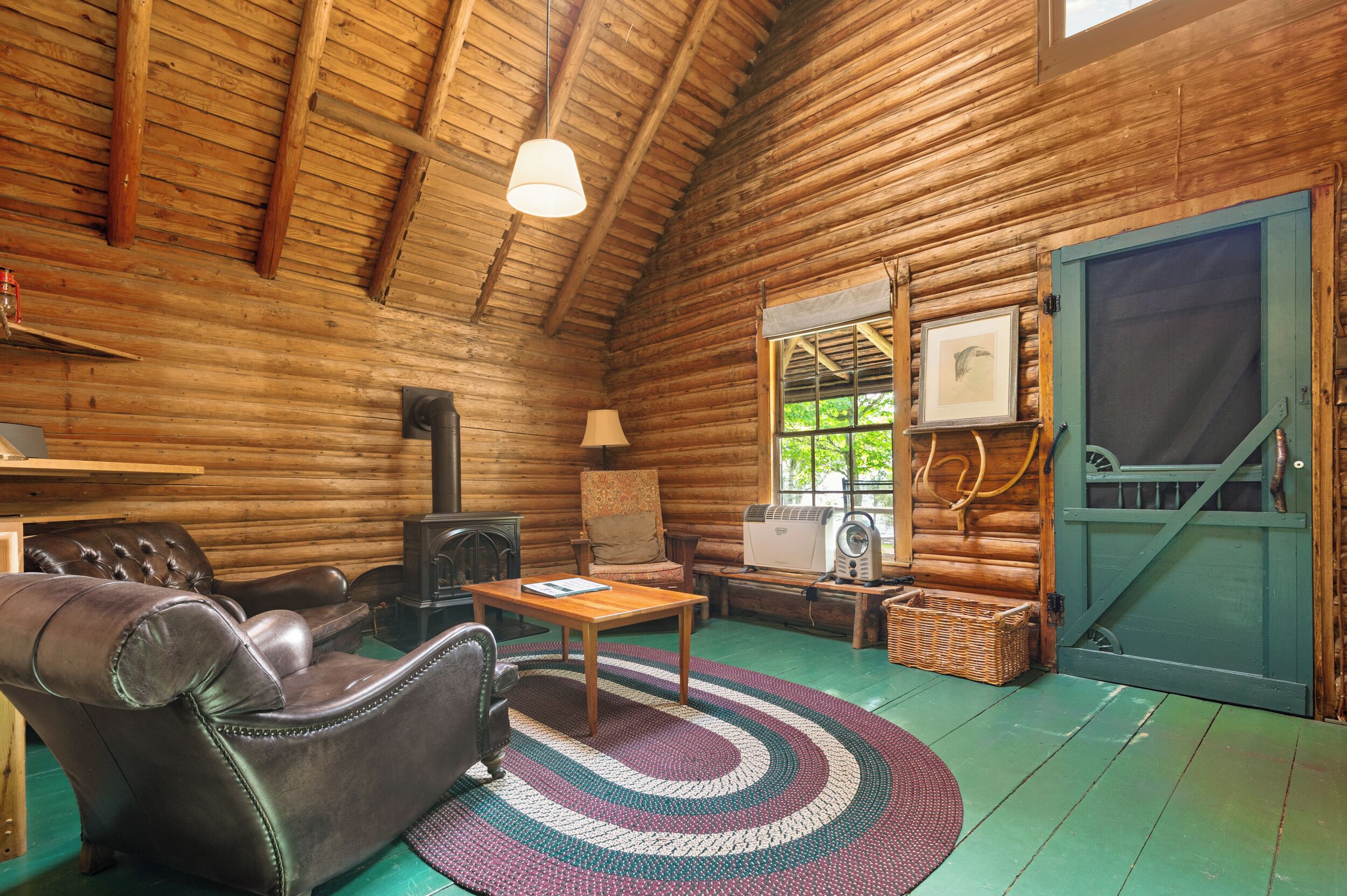 A log cabin interior with a leather sofa, a wooden table, a fireplace, and a large window.