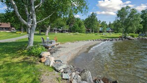 A lakeside property with a grassy area, a dock, and several houses in the background. - LuckyStryk - Classic Barker Loop Camp on Mooselookmeguntic (Oquossoc)