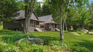 A log cabin with a porch and a deck, surrounded by lush greenery and trees. - LuckyStryk - Classic Barker Loop Camp on Mooselookmeguntic (Oquossoc)