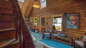 A log cabin interior with a loft bedroom, wooden walls, and a large window. - LuckyStryk - Classic Barker Loop Camp on Mooselookmeguntic (Oquossoc)