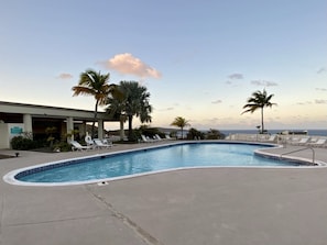 A swimming pool with lounge chairs and palm trees, surrounded by a building with a view of the ocean.