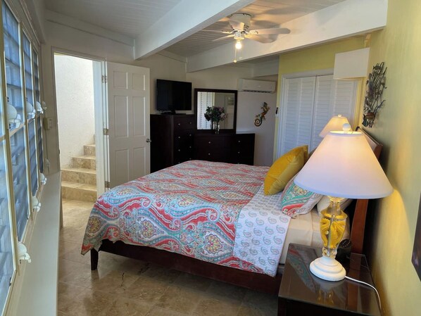 A bedroom with a bed, a nightstand, a lamp, a dresser, a television, and a window with blinds.