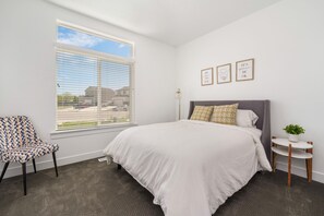 Une chambre avec une grande fenêtre, un lit recouvert de draps blancs, une chaise à motifs et deux tableaux accrochés au mur.