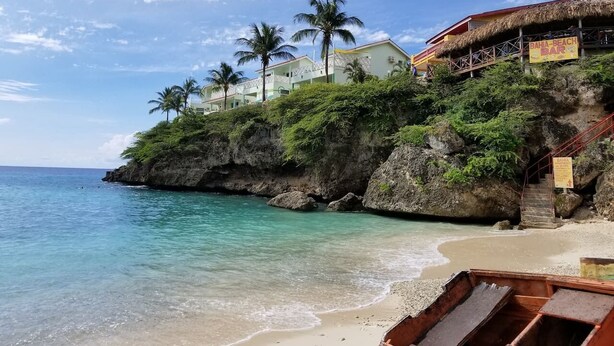 Uma praia com águas turquesa cristalinas, areal arenoso e um edifício com telhado de palha, com a placa 'BAHA BEACH BAR'.