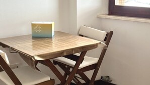 A room with a wooden table and white cushioned chairs, a window with blinds, and a wall-mounted shelf. - S296 - Sirolo, studio flat in the center with sea view (Sirolo)