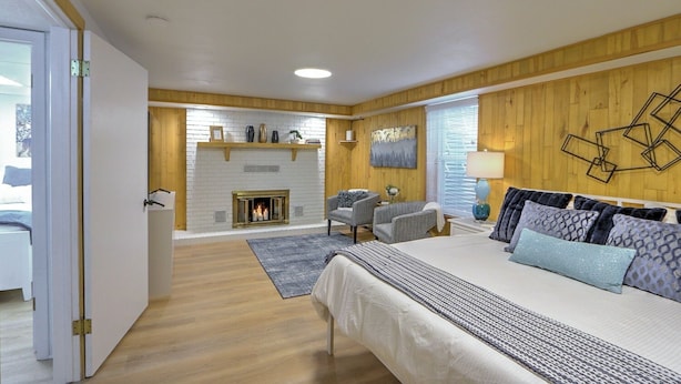 Un dormitorio con chimenea, cama con almohadas estampadas, sofá y paneles de madera.