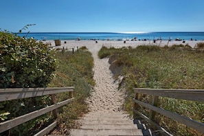 Ein Holzweg führt zu einem Sandstrand, der von einem klaren, blauen Himmel überdacht wird.