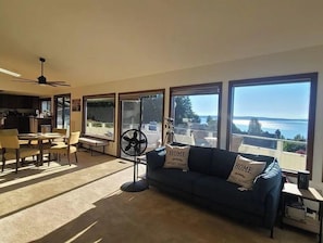 A spacious living room with a dark blue sofa, two windows with ocean views, a ceiling fan, and a dining area with chairs and a table.